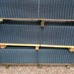 Stos szarych, metalowych paneli ogrodzeniowych na drewnianych paletach, zabezpieczonych taśmą, ułożonych na żwirowym podłożu.