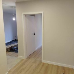 Krystian Rakoczy - Wnętrze mieszkania po remoncie: widok na nową, białą, częściowo otwartą drzwi wewnętrzne z czarną klamką, panele podłogowe w odcieniach drewna, pomalowane na jasno ściany oraz fragment...