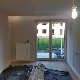 Krystian Rakoczy - Pokój w trakcie prac wykończeniowych: podłoga zabezpieczona folią, listwy przypodłogowe przy ścianie, widoczne wiadro i puszki po farbach za oknem balkonowym.
