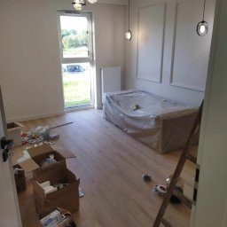 Krystian Rakoczy - Pokój w trakcie odświeżania: zabezpieczone meble folią, kartony z materiałami budowlanymi na podłodze, drewniana drabina oparta o ścianę, nowoczesne lampy sufitowe.