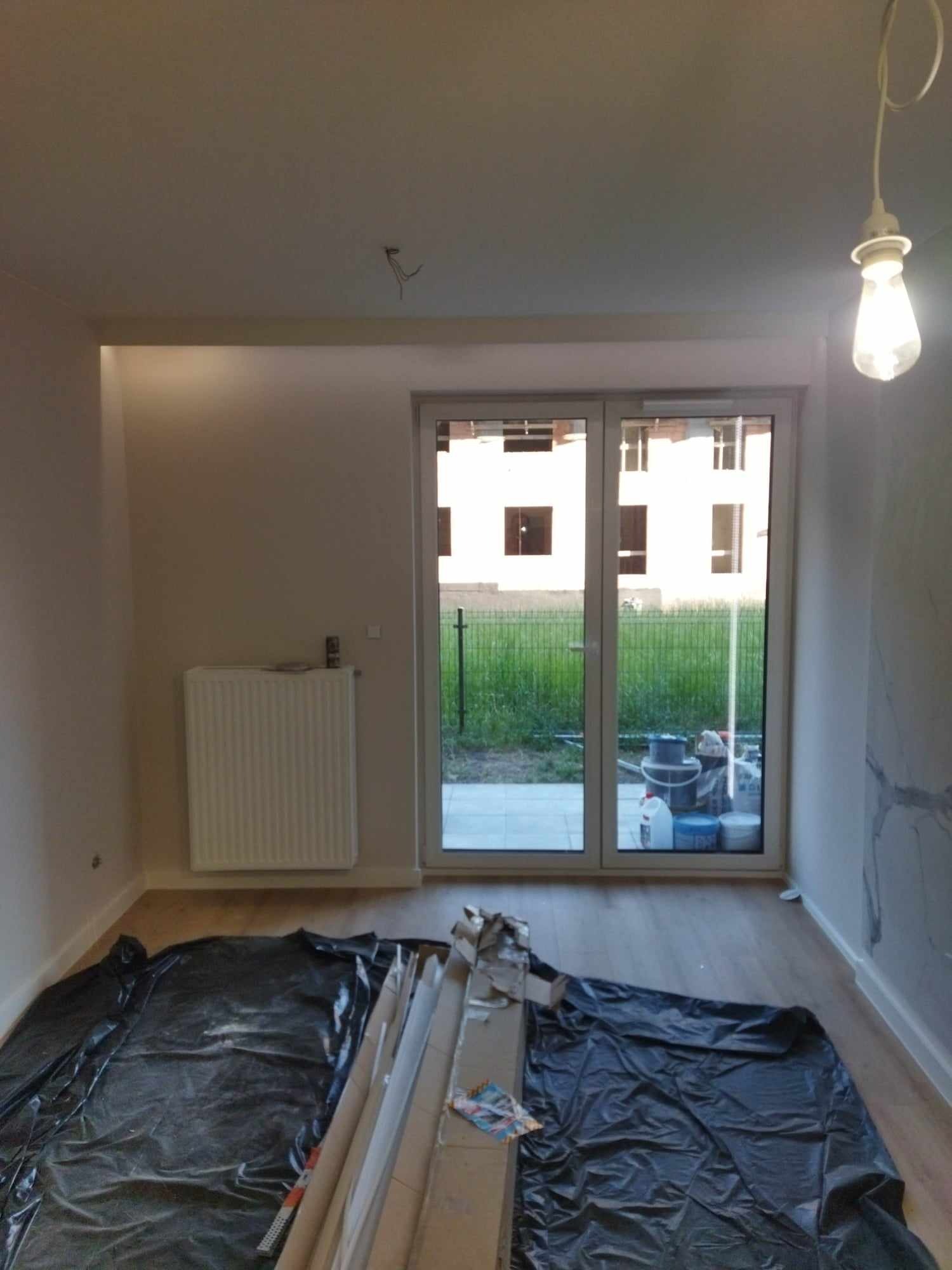 Pokój w trakcie prac wykończeniowych: podłoga zabezpieczona folią, listwy przypodłogowe przy ścianie, widoczne wiadro i puszki po farbach za oknem balkonowym.