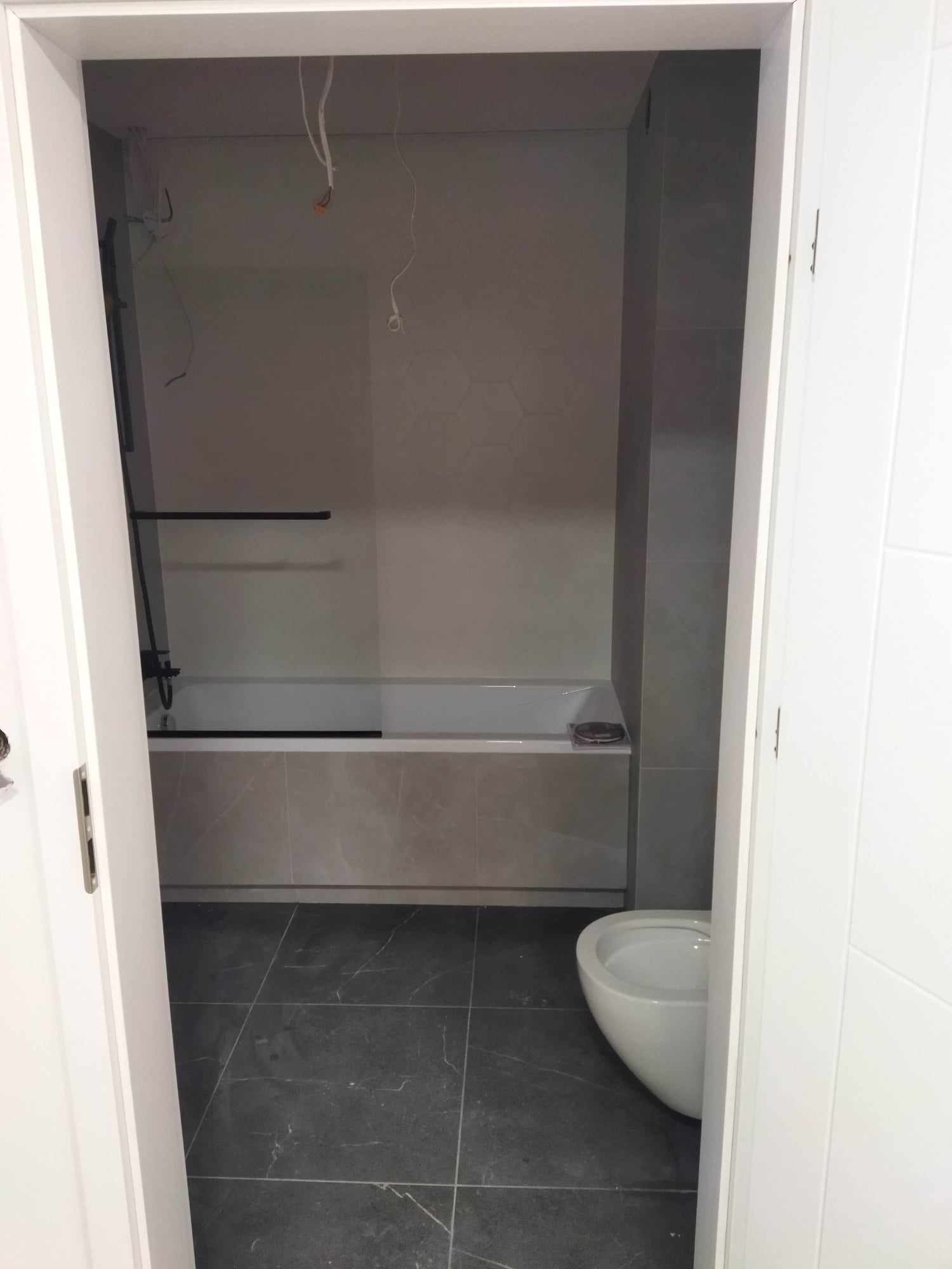 Widok przez otwarte drzwi na remontowaną łazienkę z szarymi płytkami podłogowymi i ściennymi, białą wanną z czarną armaturą i wiszącą toaletą; widoczne wystające kable.