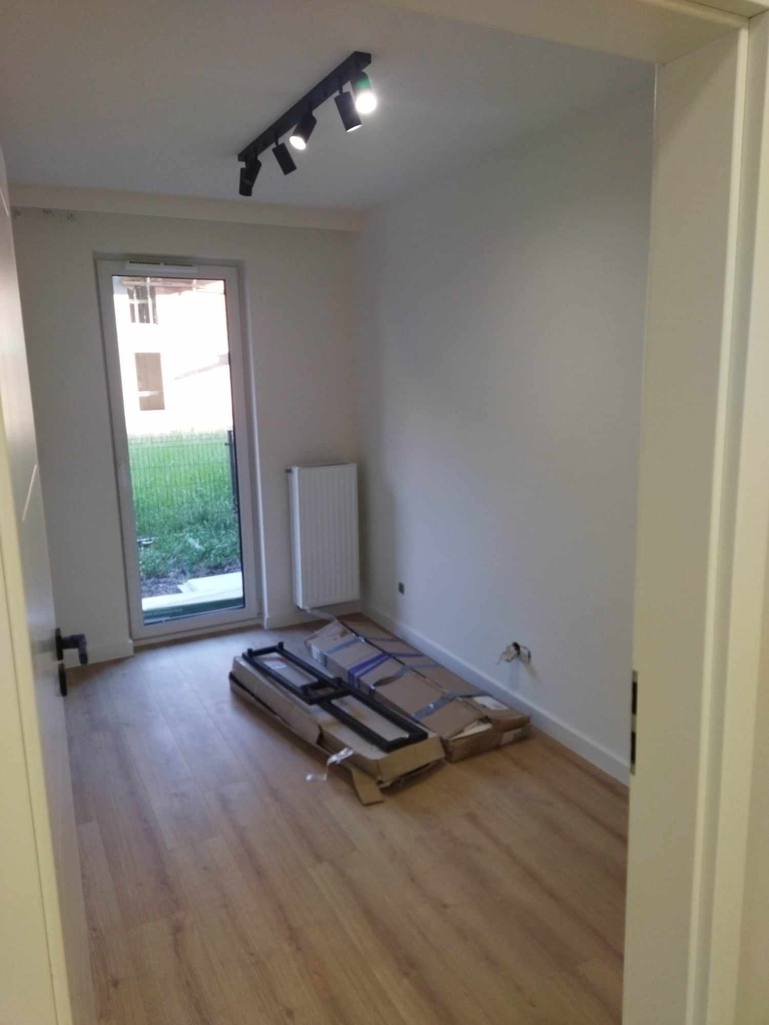 Pusty pokój z drewnianą podłogą, białymi ścianami i zamontowanym oświetleniem sufitowym, z rozpakowanymi drzwiami w kartonowych opakowaniach, czekającymi na montaż.