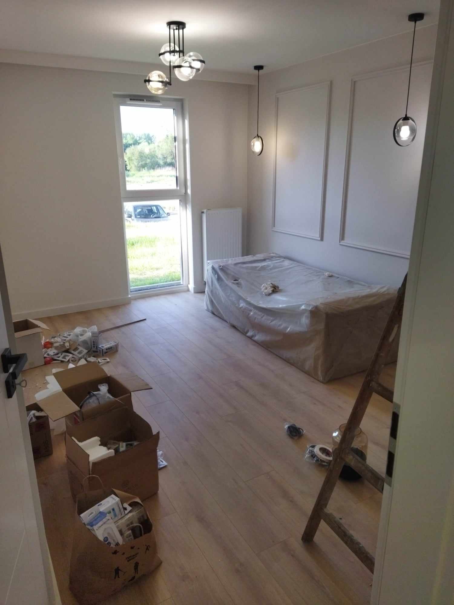 Pokój w trakcie odświeżania: zabezpieczone meble folią, kartony z materiałami budowlanymi na podłodze, drewniana drabina oparta o ścianę, nowoczesne lampy sufitowe.