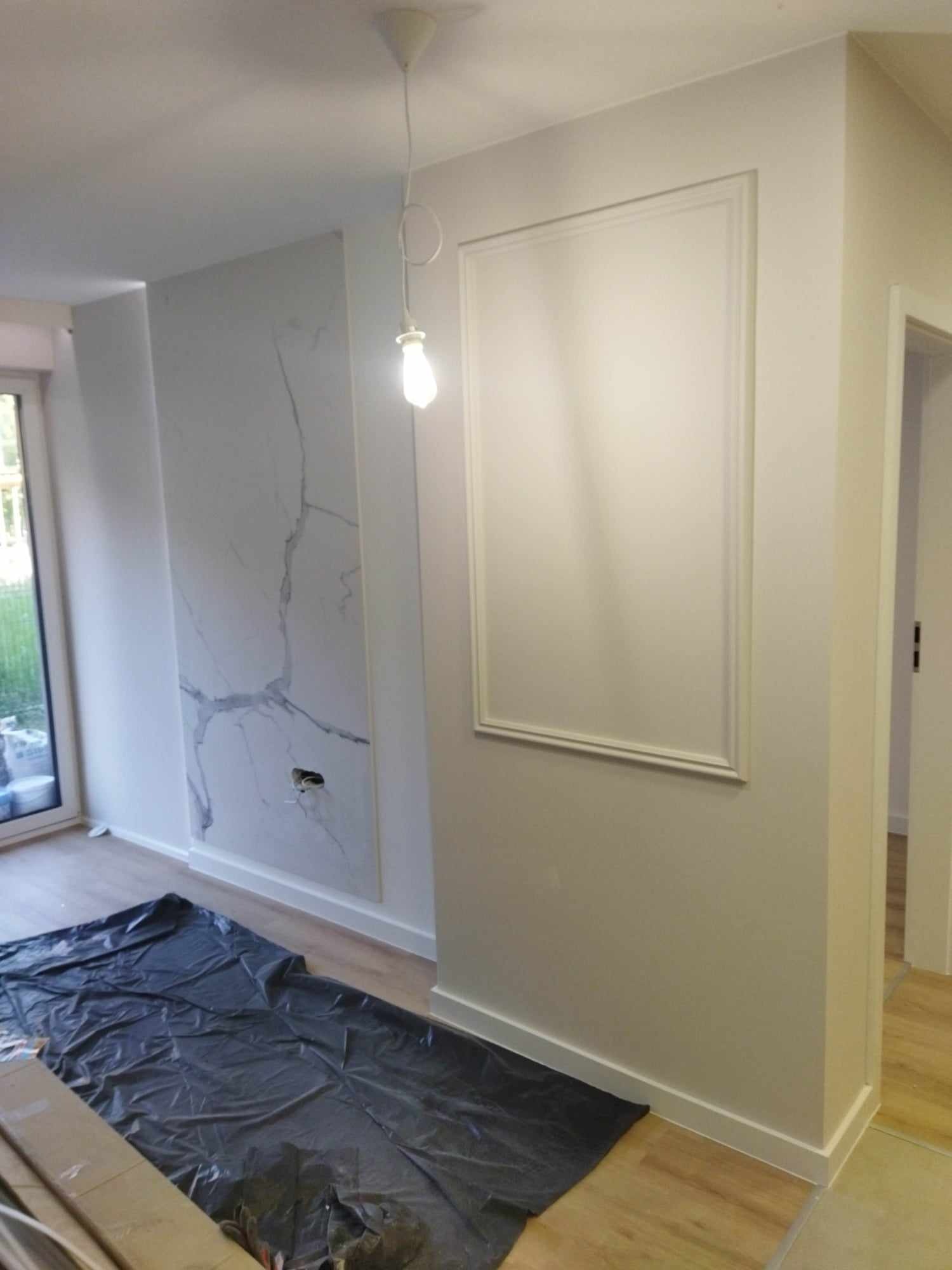 Pokój w trakcie odświeżania: ściana z imitacją marmuru, dekoracyjna rama na ścianie, podłoga zabezpieczona folią malarską, surowa żarówka zwisająca z sufitu.