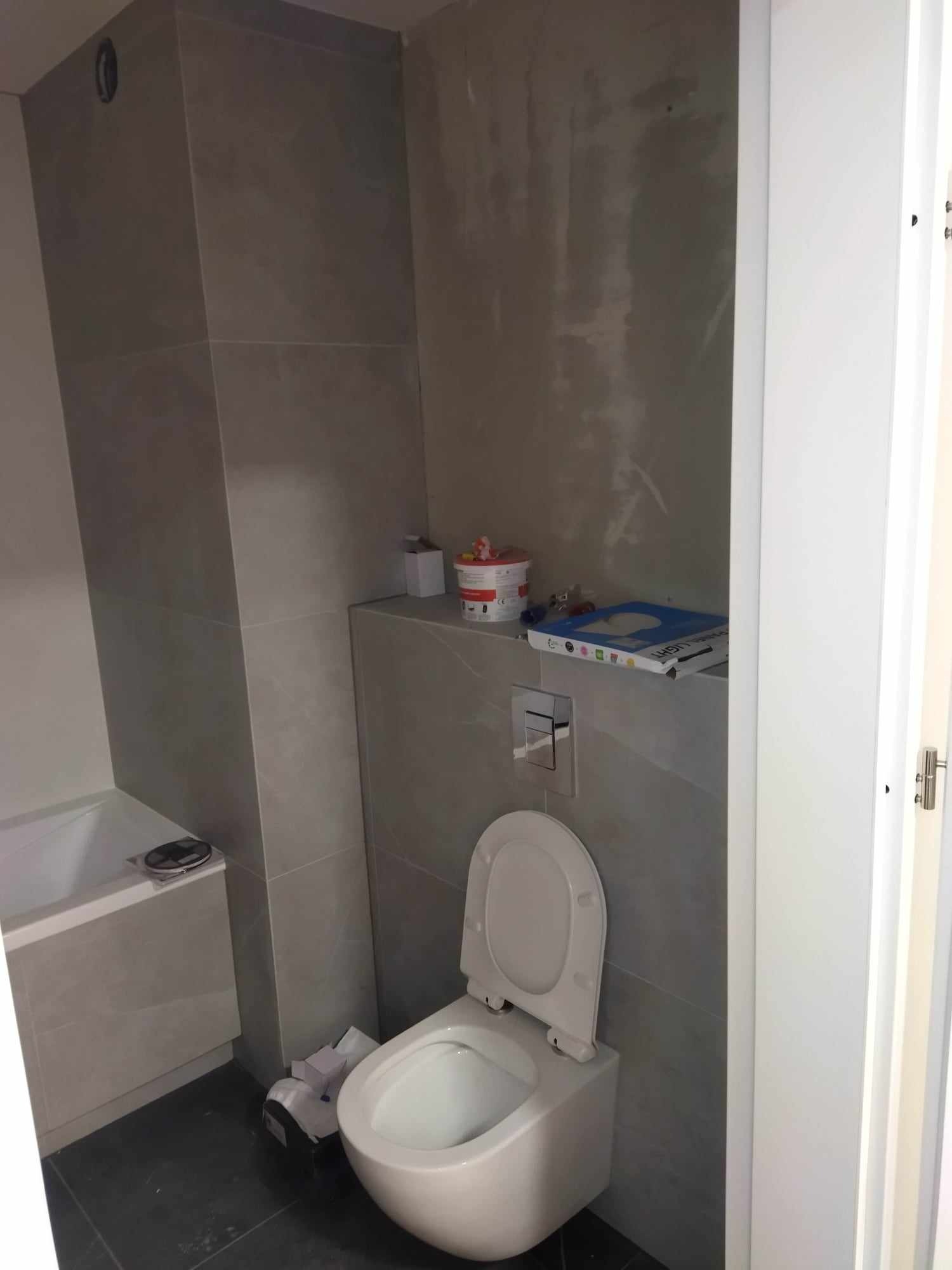 Nowa toaleta wisząca z otwartą klapą w łazience z szarymi płytkami, widoczne narzędzia i materiały budowlane na półce nad toaletą.