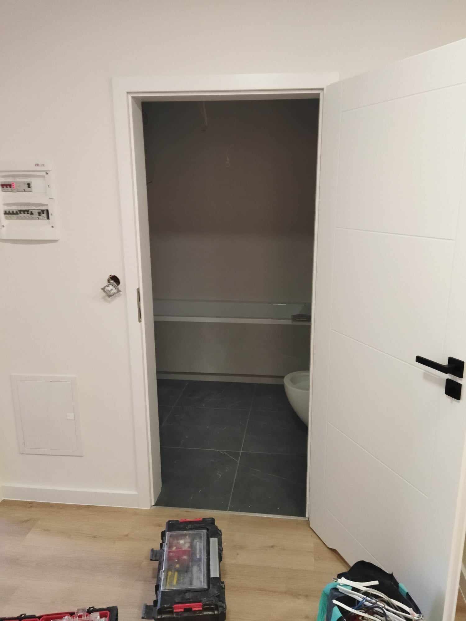 Otwarta biała, nowoczesna drzwi łazienkowa z czarną klamką, widok na wnętrze łazienki z wanną i toaletą, skrzynka z narzędziami na podłodze.