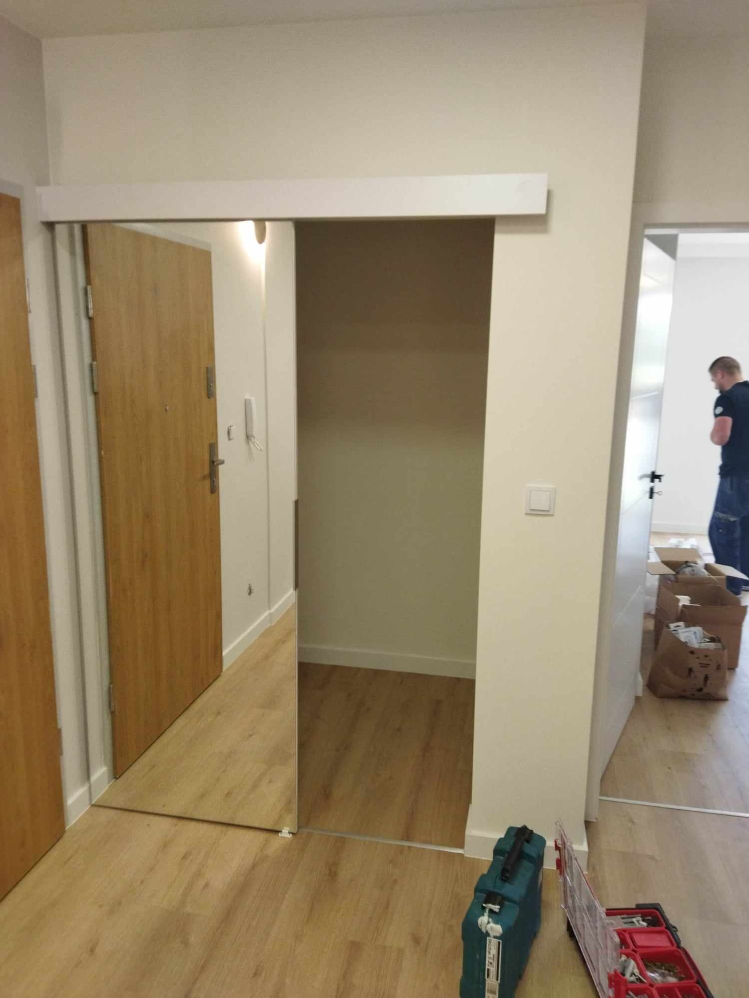 Montaż przesuwnych drzwi lustrzanych w przedpokoju, widoczne narzędzia i części montażowe na podłodze.