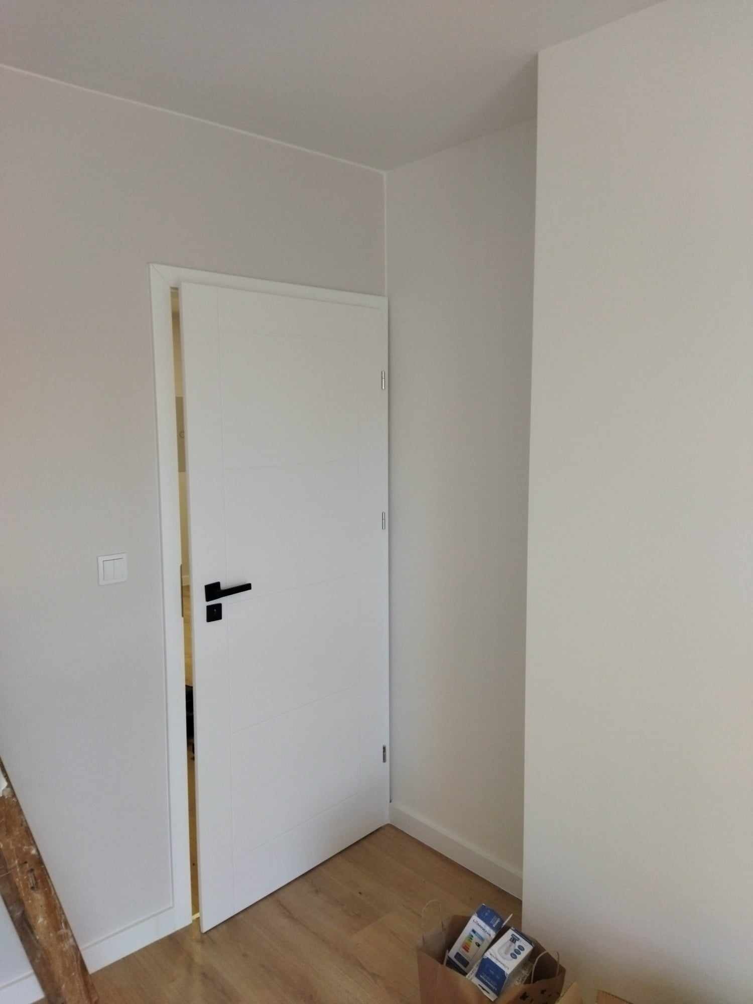 Nowo zamontowane, białe drzwi wewnętrzne z czarną klamką, widoczne na tle świeżo pomalowanej ściany w odcieniach szarości, z fragmentem podłogi wyłożonej panelami oraz kartonem z żarówkami w rogu.
