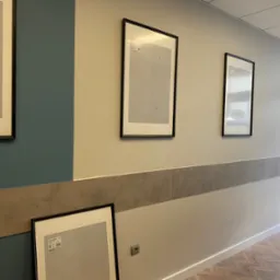 Korytarz z pomalowanymi ścianami w dwóch odcieniach, z zamontowanymi listwami dekoracyjnymi i trzema pustymi ramami na zdjęcia, z czego jedna oparta o ścianę.