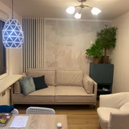 Jasny salon z beżową sofą, białym fotelem, stolikiem i wiszącą lampą o geometrycznym wzorze, ściana ozdobiona tapetą z motywem drzew.