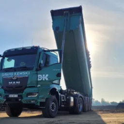 Zielona wywrotka MAN firmy David Kemski z podniesioną skrzynią na placu budowy, w tle hałdy ziemi i błękitne niebo z śladem po samolocie.