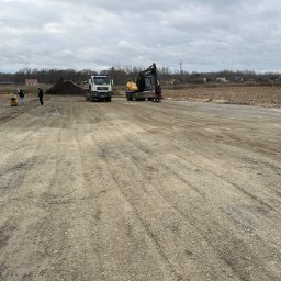 DK SPÓŁKA Z OGRANICZONĄ ODPOWIEDZIALNOŚCIĄ - Przygotowanie terenu pod budowę: ciężarówka MAN wyładowuje materiał obok koparki Volvo, w tle dwóch pracowników obsługuje zagęszczarkę gruntu.