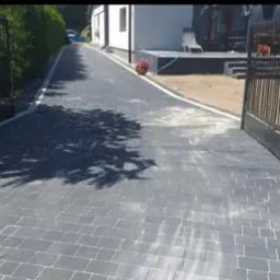 Ułożona szara nawierzchnia z kostki brukowej prowadząca od bramy wjazdowej do domu, z widocznymi cieniami drzew.