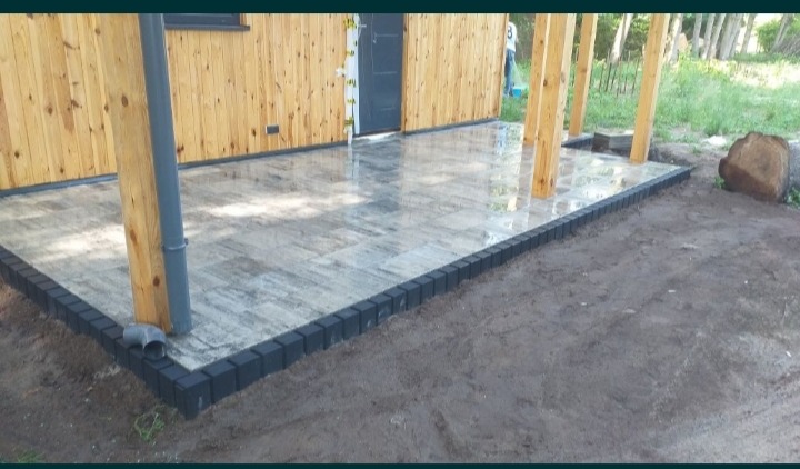 Ułożona, szaro-beżowa kostka betonowa na tarasie przylegającym do drewnianego budynku z widocznymi słupami, obrzeża z ciemnej kostki, rura spustowa przy słupie.