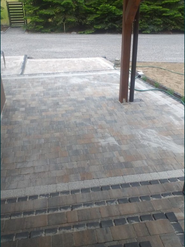 Wykończona nawierzchnia z kostki brukowej o nieregularnym wzorze i obramowaniu z ciemniejszych kostek, widok z perspektywy wejścia na posesję.