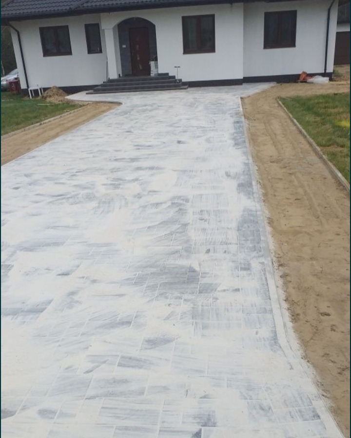 Ułożona szara kostka na podjeździe przed domem, z widocznymi śladami białego piasku lub cementu na powierzchni, sugerując prace wykończeniowe.