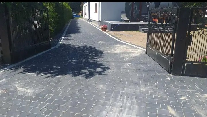 Ułożona szara nawierzchnia z kostki brukowej prowadząca od bramy wjazdowej do domu, z widocznymi cieniami drzew.