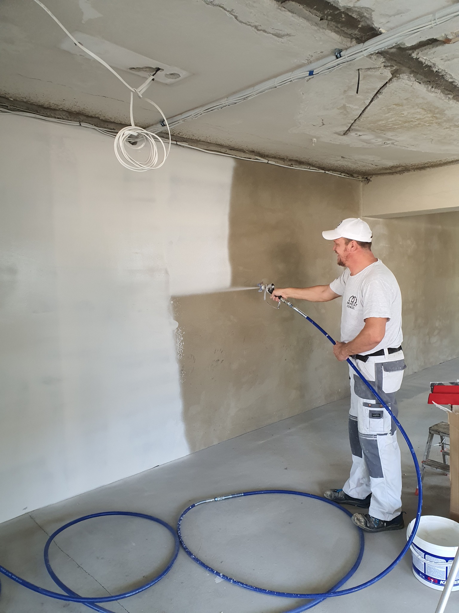 Mężczyzna w kombinezonie malarskim, z uśmiechem, natryskuje białą farbę na ścianę w surowym wnętrzu, widoczny niepomalowany fragment ściany, niebieski wąż od agregatu leży na podłodze.