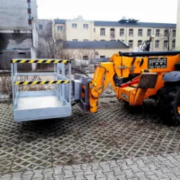 Żółta ładowarka teleskopowa JCB z zamontowanym koszem roboczym na tle miejskiej zabudowy, stojąca na ażurowej kostce brukowej.