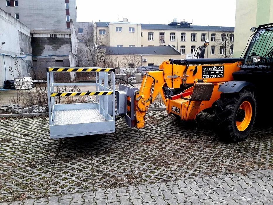 Żółta ładowarka teleskopowa JCB z zamontowanym koszem roboczym na tle miejskiej zabudowy, stojąca na ażurowej kostce brukowej.