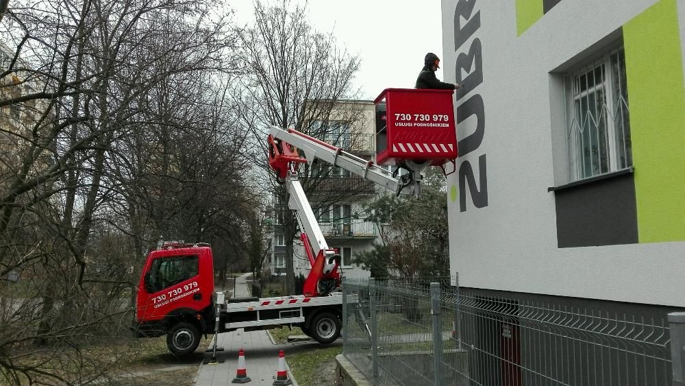 Czerwony podnośnik koszowy na pojeździe z numerem telefonu, pracownik w koszu na tle budynku z napisem ZUBR, widok z poziomu ulicy.