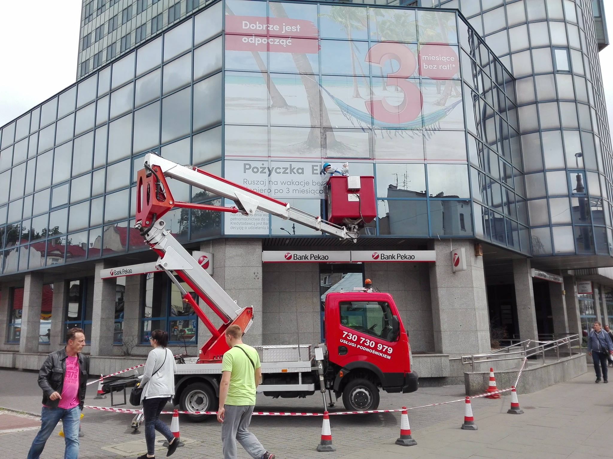 Czerwony podnośnik koszowy na tle budynku Banku Pekao, z pracownikiem w koszu na wysokości, prawdopodobnie montującym lub czyszczącym reklamę na szklanej fasadzie.