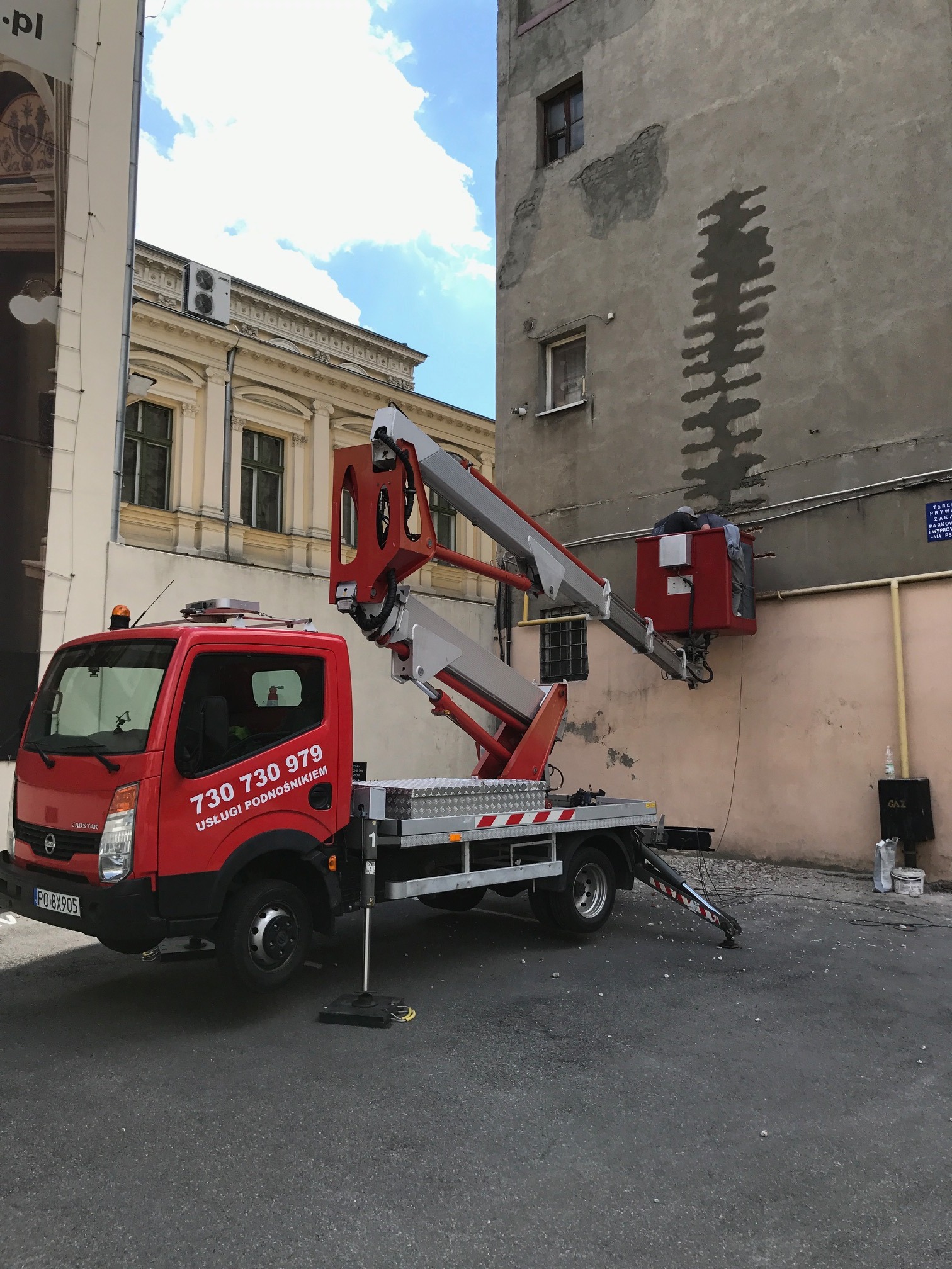 Czerwony podnośnik koszowy na podwoziu samochodu ciężarowego, z wysięgnikiem sięgającym do ściany kamienicy z widocznymi śladami czyszczenia, pracownik w koszu, w tle budynek z klimatyzatorami.