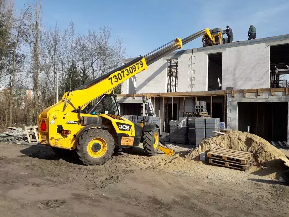 Żółta ładowarka teleskopowa JCB 535-140 z wysuniętym ramieniem podnosi materiały budowlane na budowę domu jednorodzinnego, na dachu dwóch robotników w ciemnych ubraniach.