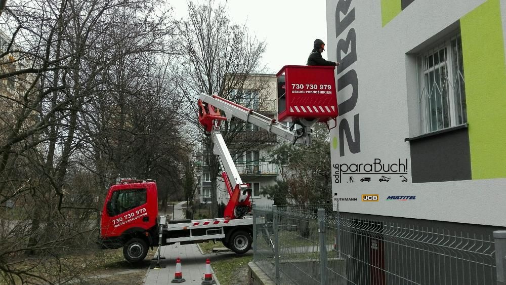 Czerwony podnośnik koszowy z numerem telefonu na tle budynku z reklamą firmy ZUBR, pracownik w koszu podnośnika podczas pracy.