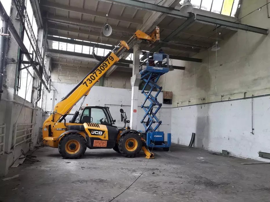 Żółta ładowarka teleskopowa JCB z wysięgnikiem podtrzymującym niebieski podnośnik nożycowy wewnątrz hali z betonową posadzką i ścianami.