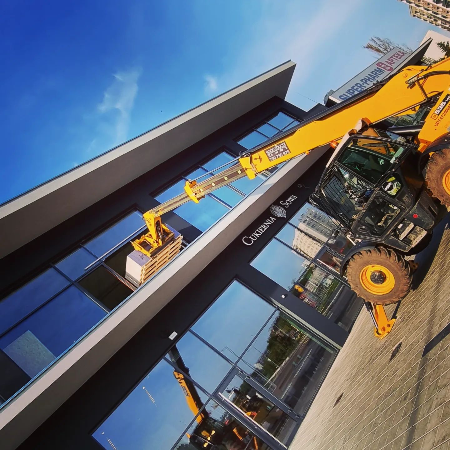 Żółta maszyna teleskopowa z paletą przed oknem budynku z szyldami 'Cukiernia Sowa' i 'Super-Pharm Apteka', widok z dołu na błękitne niebo.