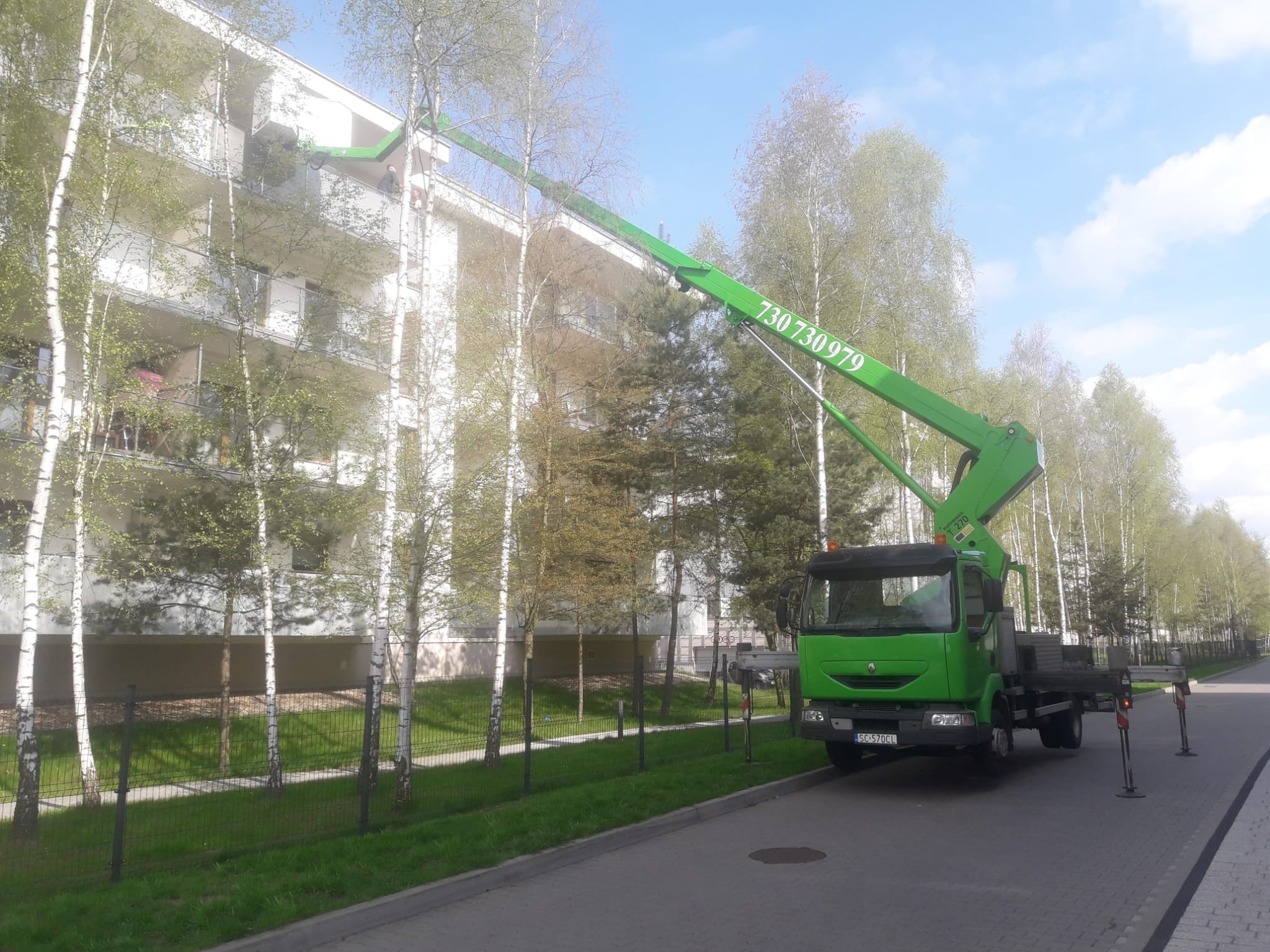 Zielony podnośnik koszowy na ulicy, z wysięgnikiem sięgającym do balkonu budynku mieszkalnego, widoczne brzozy w otoczeniu.