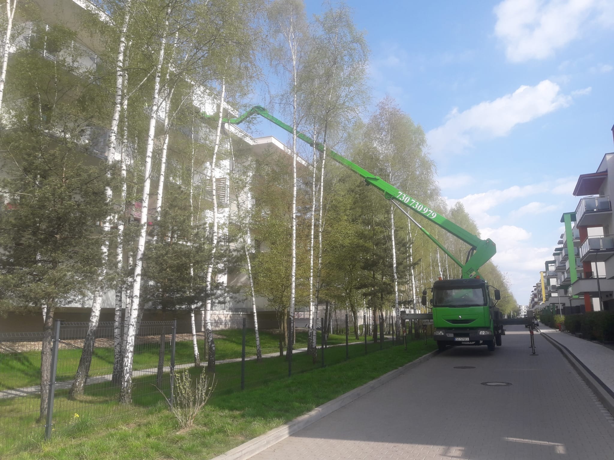 Zielony podnośnik koszowy na chodniku, wysięgnik sięga do rynny budynku mieszkalnego z białymi balkonami, obok rosną brzozy.