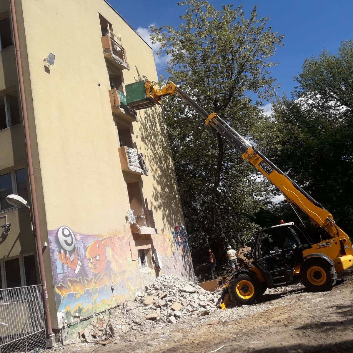 Żółta ładowarka teleskopowa z zielonym kontenerem na wysięgniku, pracująca przy rozbiórce balkonu budynku z graffiti. Gruz na ziemi, widoczni pracownicy w tle.