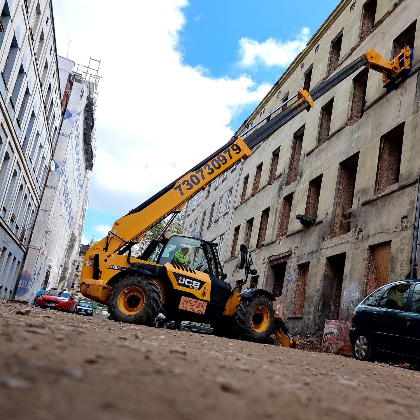 Żółta ładowarka teleskopowa JCB z wysięgnikiem sięgającym do okien starego, ceglanego budynku w trakcie prac remontowych; widok z dołu, na pierwszym planie gruz i zaparkowane samochody