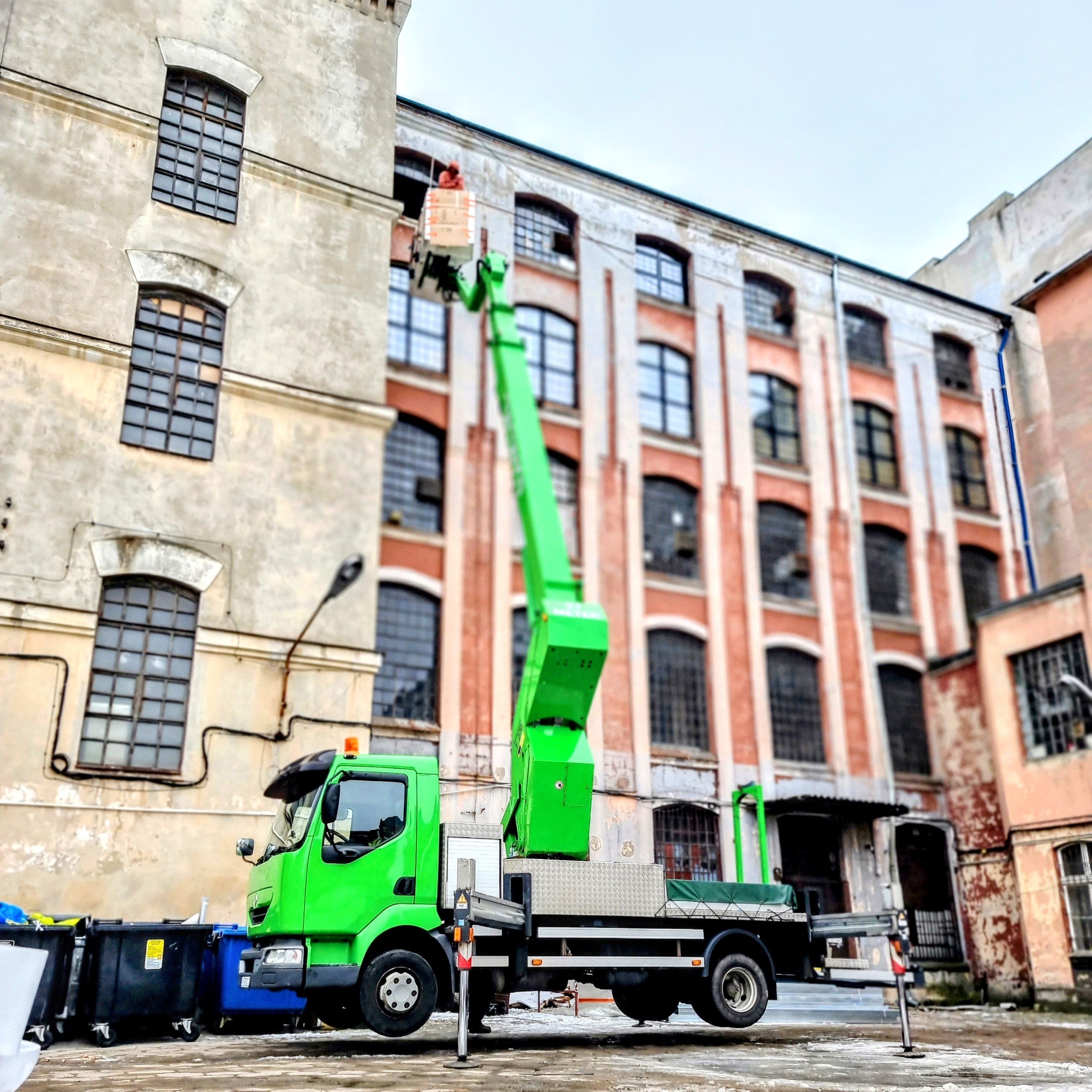 Jasnozielony podnośnik koszowy na tle starych budynków z cegły i betonu, osoba w koszu wykonuje prace na wysokości.