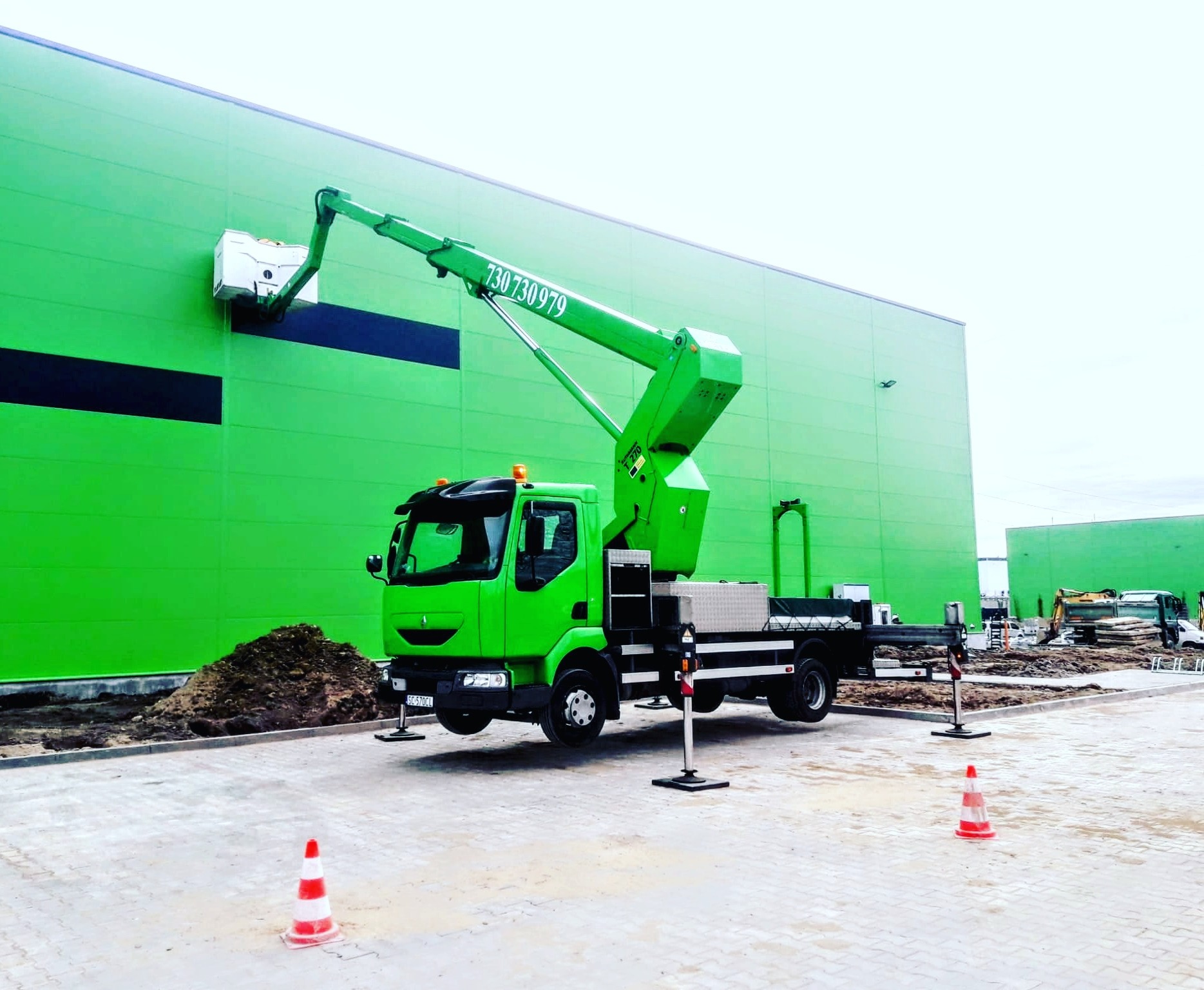 Zielony podnośnik koszowy na tle zielonej hali, z wysięgnikiem skierowanym ku górze, podczas prac na elewacji, widoczne pachołki ostrzegawcze i zaplecze budowy.