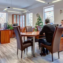 Jasne biuro z drewnianymi meblami, pracownikami przy biurkach i choinką w tle, widziane z perspektywy klienta siedzącego przy okrągłym stole.