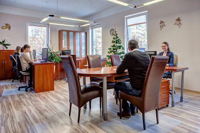 Jasne biuro z drewnianymi meblami, pracownikami przy biurkach i choinką w tle, widziane z perspektywy klienta siedzącego przy okrągłym stole.