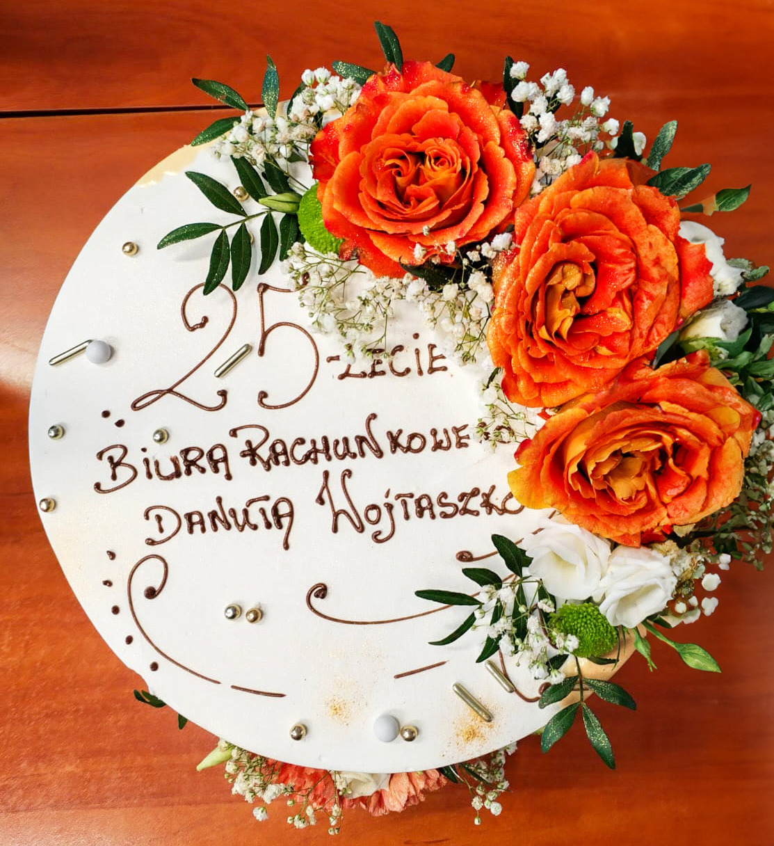 Tort z okazji 25-lecia Biura Rachunkowego Danuta Wojtaszko, udekorowany pomarańczowymi różami, białymi kwiatami i zielonymi listkami, na drewnianym stole.