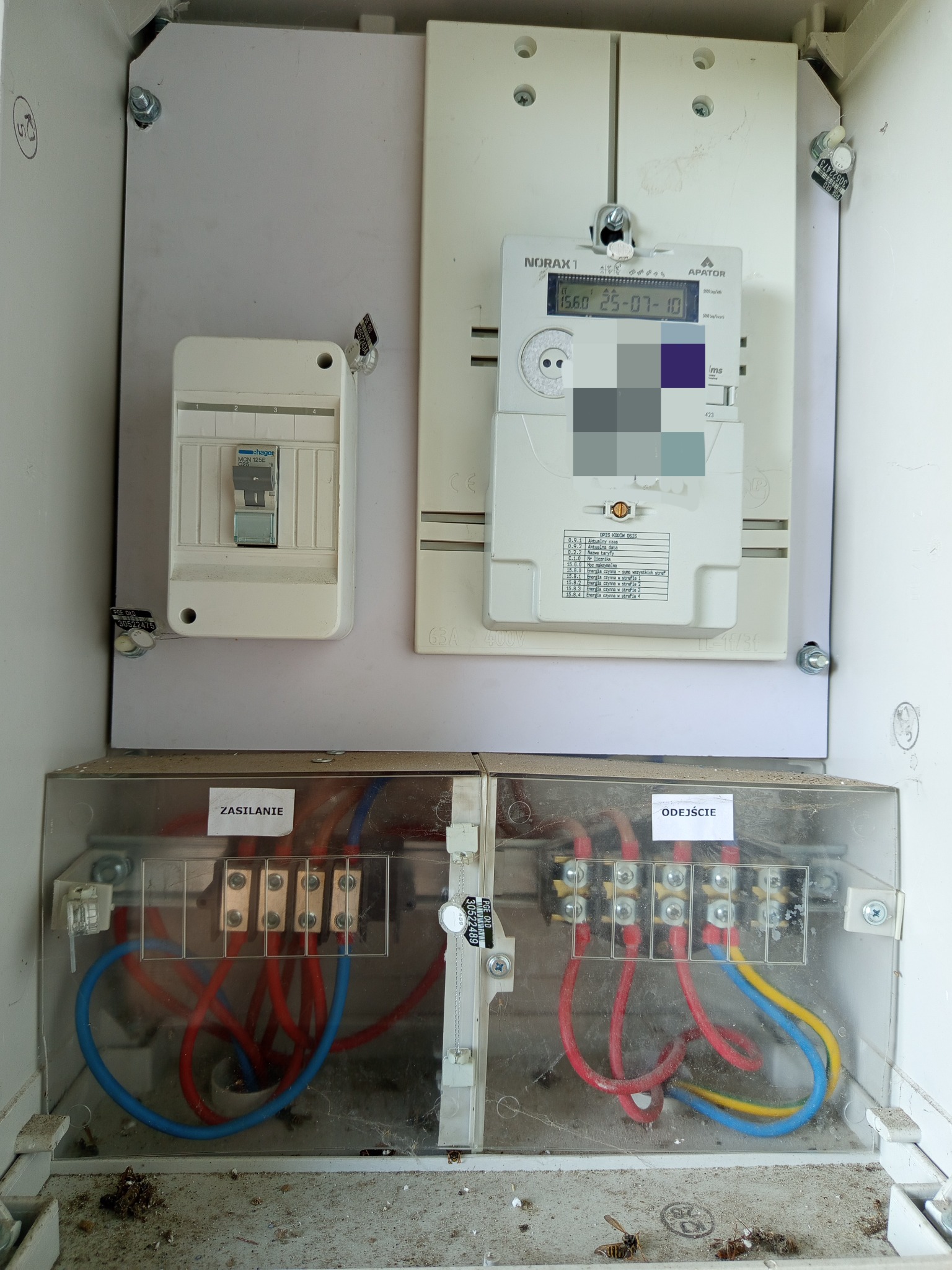 Rozdzielnia elektryczna z licznikiem, bezpiecznikiem i okablowaniem. Widoczne oznaczenia 'Zasilanie' i 'Odejście'. Elementy zakurzone, co sugeruje długotrwałe użytkowanie.