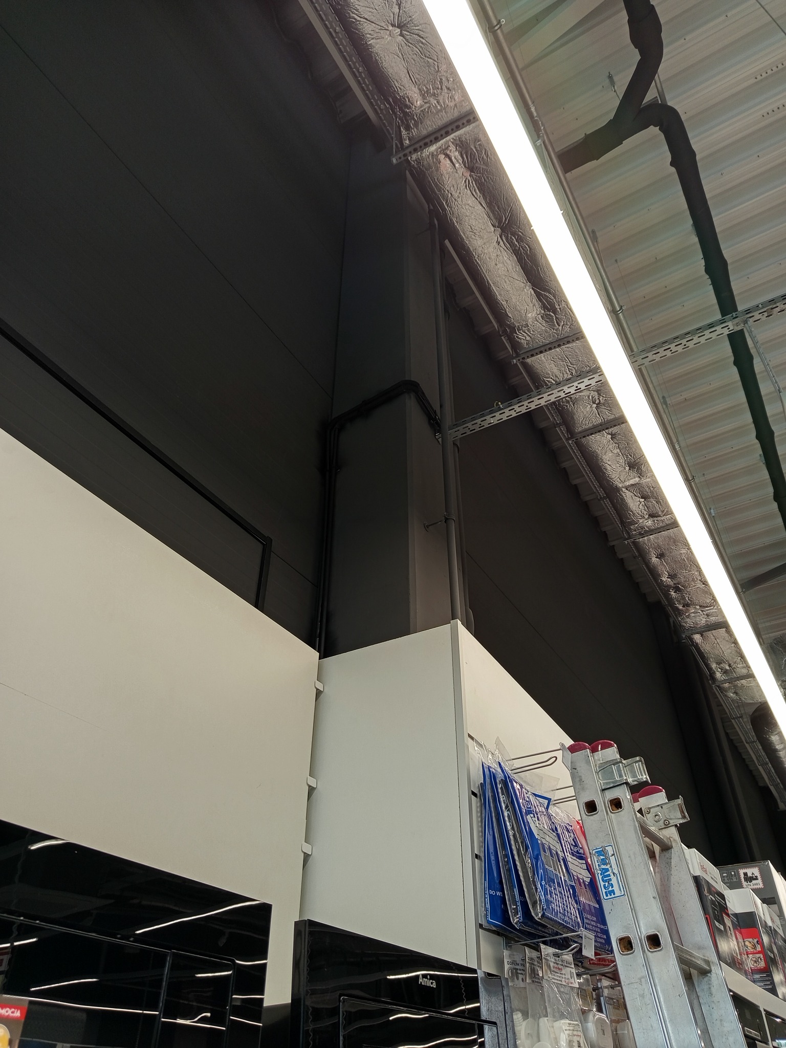 Wnętrze sklepu z oświetleniem: widoczna instalacja elektryczna pod sufitem, rury, kable i lampa fluorescencyjna. W tle regały z produktami i drabina.