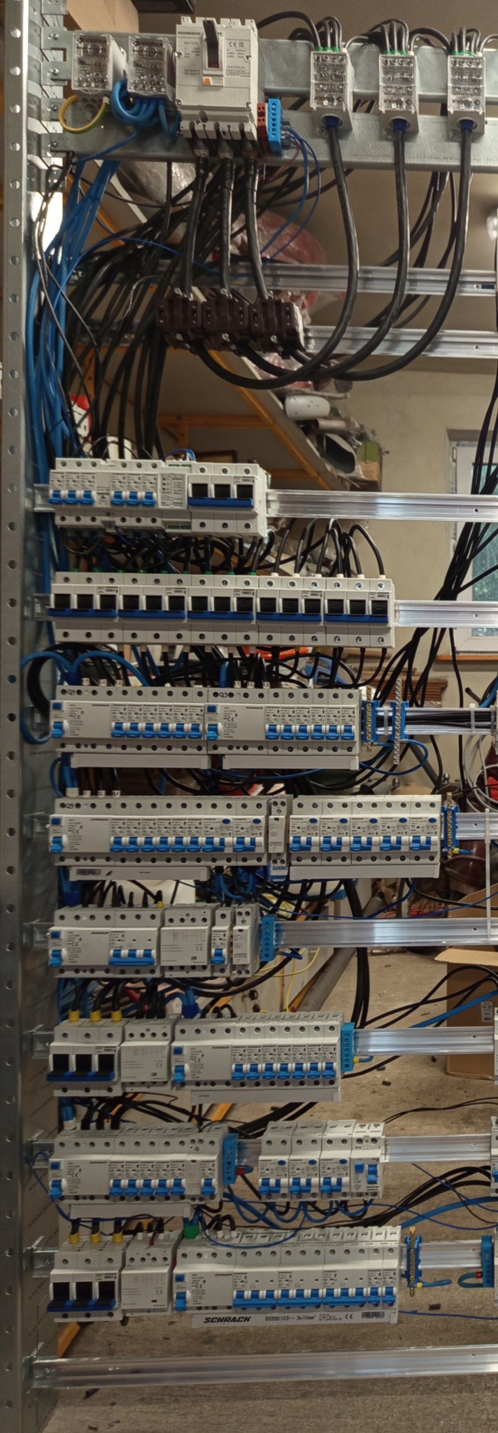 Rozdzielnia elektryczna z licznymi zabezpieczeniami i przewodami, zamontowana na metalowej konstrukcji. Widoczne bezpieczniki i kable w kolorach czarnym i niebieskim.