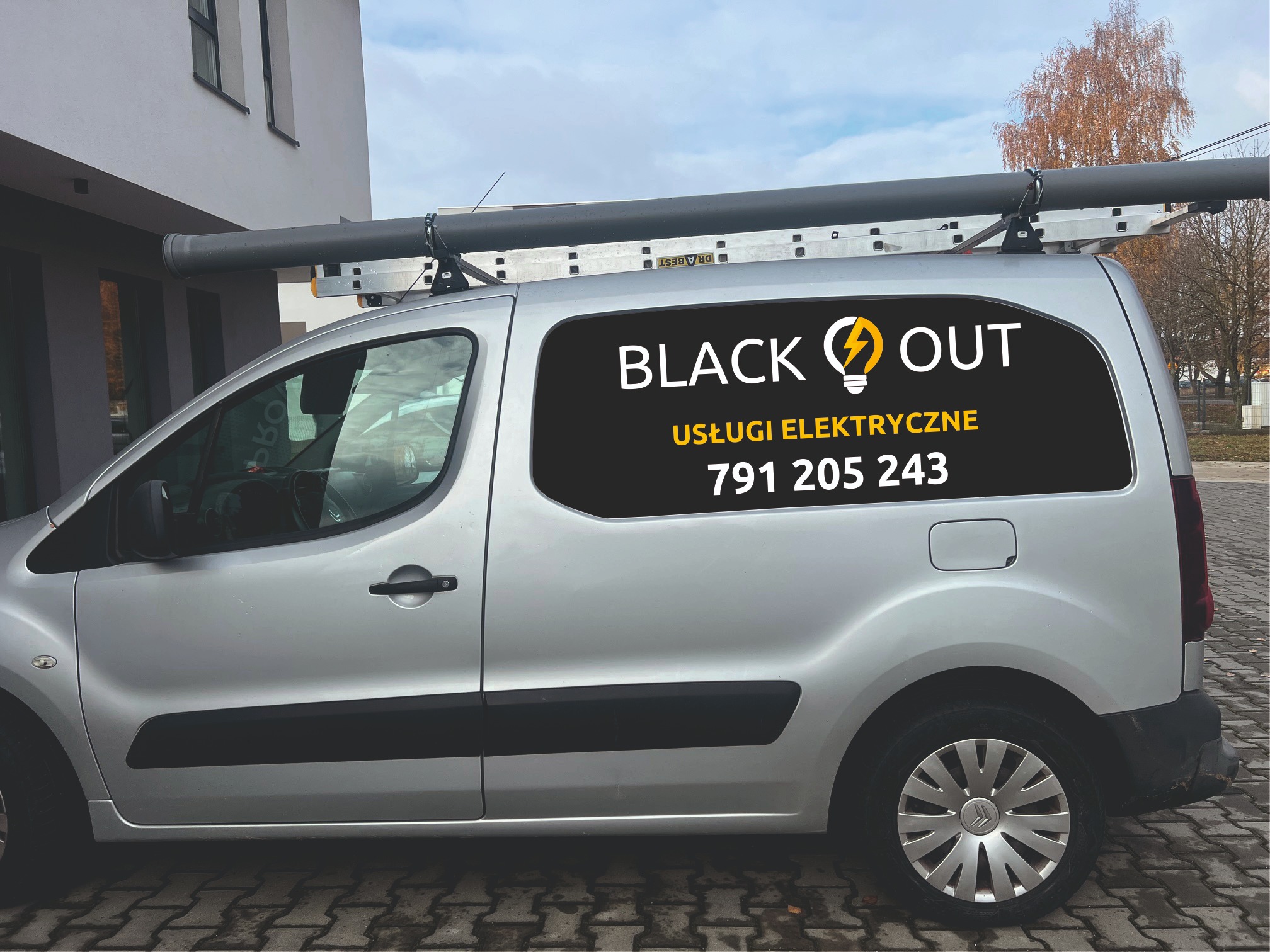 Srebrny samochód dostawczy z drabiną na dachu i reklamą usług elektrycznych 'BLACK OUT' z numerem telefonu na boku, zaparkowany na brukowanej ulicy.