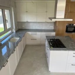 Jasna kuchnia z białymi szafkami i szarym blatem z kamienia naturalnego, wyspa kuchenna z płytą gazową, okno z widokiem na ogród.