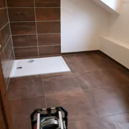 Nowa kabina prysznicowa z brązowymi płytkami, widoczne przyłącza wodne i lampa budowlana na podłodze.