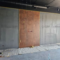 RAF-REMO Usługi remontowo-budowlane Rafał Nizio - Ściana w trakcie prac wykończeniowych: szare ściany po bokach, centralnie brązowe płytki, zabezpieczona podłoga, widoczne instalacje elektryczne.