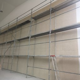 RAF-REMO Usługi remontowo-budowlane Rafał Nizio - Montaż płyt na ścianie z użyciem rusztowania. Widoczne płyty w odcieniach beżu, metalowe rusztowanie i drabina. Oświetlenie sufitowe.