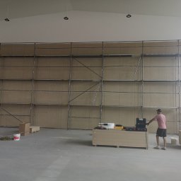 RAF-REMO Usługi remontowo-budowlane Rafał Nizio - Montaż płyt na ścianie w dużym pomieszczeniu. Widoczne rusztowania, narzędzia i pracownik w trakcie pracy. Oświetlenie sufitowe.